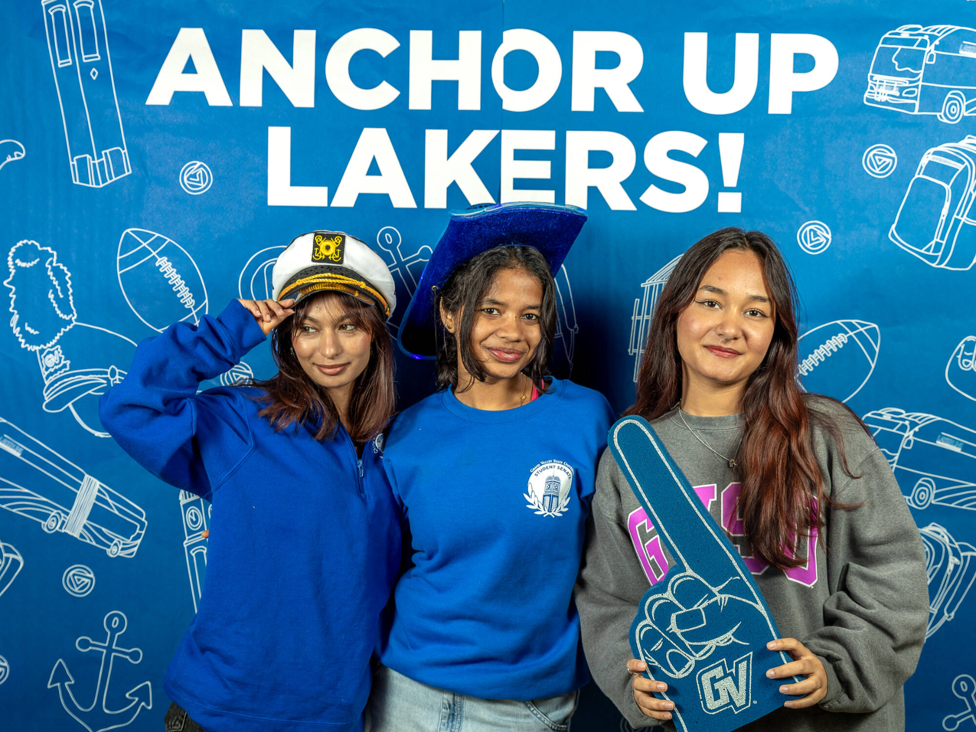 three individuals, one on the left wearing a sailor hat, one in the middle wearing a blue cowboy hat, one on the right holding up a foam finger
